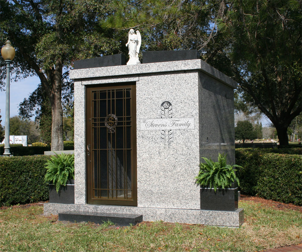Tall light grey stone mausoleum with angel statue on top and a single metal door. Also has square metal vases at the base on each side.